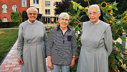 Drei Frauen, davon zwei in Ordenstracht, stehen in einem Garten.