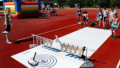 Kinder stehen auf einem Sportplatz und spielen auf einer Kunststoff-Bahn Eisstock-Schießen.