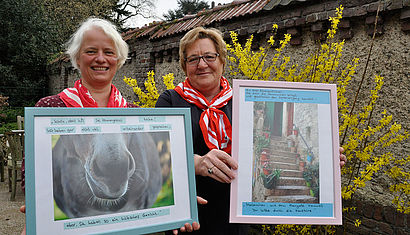 Martina Zimmer und Maria Haaken stehen in einem Garten. Sie halten zwei gerahmte Fotos in die Kamera.