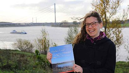 Eine Frau steht an einem Fluss und hält ein Buch in den Händen.