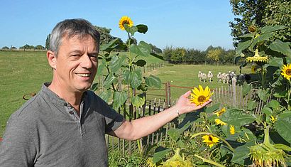Reiner Rosenberg steht in seinem Garten und hält die Blüte einer Sonnenblume. Im Hintergrund sieht man einen Deich und eine Schafherde.