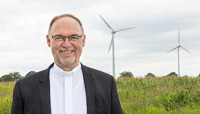 Weihbischof Rolf Lohmann steht vor einer Wiese, auf der zwei Windräder stehen.