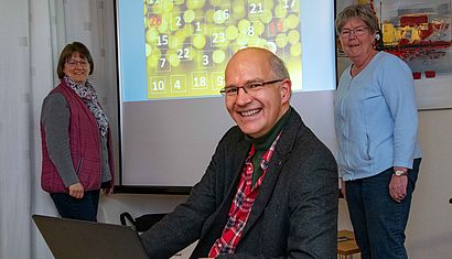 Zwei Frauen stehen neben einer Leinwand mit einem projizierten Adventskalender, davor sitzt ein Mann an einem Laptop.