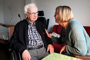 Eine Frau und ein Mann sitzen an einem Tisch. Die Frau hat die Hand auf den Arm des Mannes gelegt und spricht zu ihm. Eine Frau und ein Mann sitzen an einem Tisch. Die Frau hat die Hand auf den Arm des Mannes gelegt und spricht zu ihm.