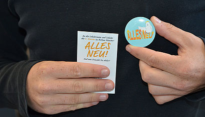 Zwei Hände halten eine kleine Karte auf der "Alles Neu" steht und einen Ansteckbutton mit dem gleichen Text.