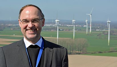 Weihbischof Lohmann steht vor mehreren Windrädern am Niederrhein.