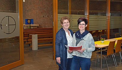 Maria Sanders und Maria Derksen, die eine rote Mappe in der Hand hält, stehen in dem Pfarrheim von Kehrum. Im Hintergrund sieht msn rechts Tische mit gelber Tischdecke, links ist der Durchgang zum Gottesdienstraum zu erkennen.