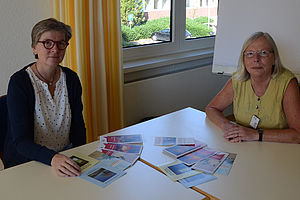 Birgit Gerhards (links) und Edith Thier sitzen nebeneinander an einem Tisch. Auf diesem liegen Flyer zu den Angeboten der Trauerbegleitung.