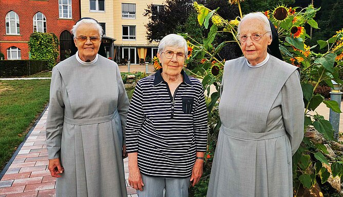 Drei Frauen, davon zwei in Ordenstracht, stehen in einem Garten.
