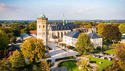 Das Bild zeigt einen großen, historischen Gebäudekomplex mit Turm im Sonnenlicht. 