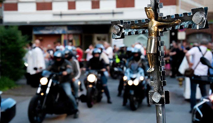 Hinter einem Vortragekreuz sind unscharf Motorräder zu erkennen.