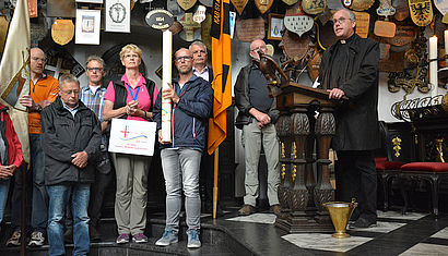 Mehrere Pilger stehen in der Kerzenkapelle in Kevelaer, eine Frau hält ein Schild zur Jubiläumswallfahrt in der Hand, daneben hält ein Mann die große Pilgerkerze. Rechts steht Pfarrer Christoph Theberath am Ambo und spricht zur Gemeinde.