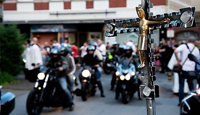 Im Vordergrund ist ein Kreuz, im Hintergrund unscharf Motorräder nebeneinander auf einer Straße.