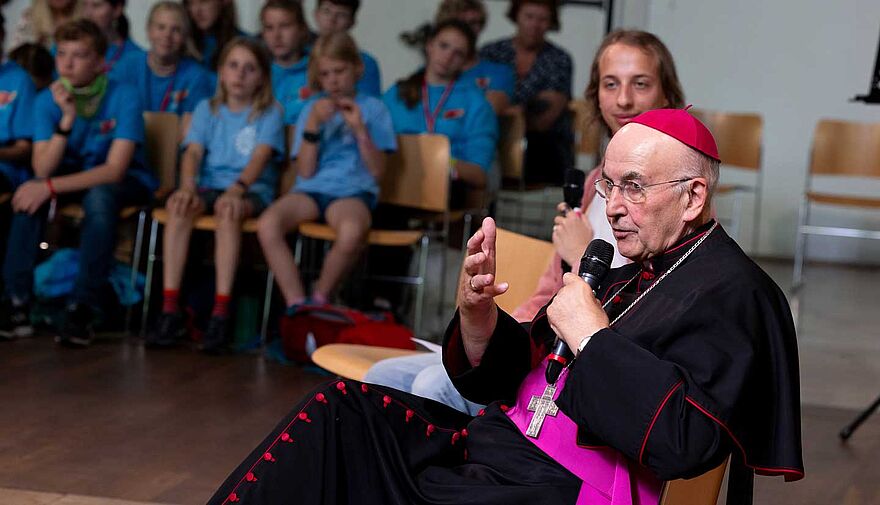 Bischof Genn sprich in ein Mikrofon, im Hintergrund sitzen Jugendliche