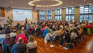 Moderatorin Laura Tietzel steht vor einer großen Leinwand und begrüßt die mehr als 160 Teilnehmenden beim Startklar!-Tag für Pfarreiräte in der Katholischen Akademie Stapelfeld. Die Besucherinnen und Besucher sitzen dicht an dicht im großen Veranstaltungssaal und verfolgen die Begrüßung.