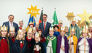  Viele Sternensinger und Sternensingerinnen mit goldenen Kronen und Umhängen in unterschiedlichen Farben, einige blau und andere grün, lila oder gelb. Dazwischen stehen die beiden Politiker. Alle lächeln fröhlich in die Kamera.: Der Segen für die nordrhein-westfälische Staatskanzlei muss auch 2026 erneuert werden. Zuletzt brachte eine Gruppe Sternsingerinnen der Gemeinde St. Josef aus Moers den Segen zum Amtssitz von Ministerpräsident Hendrik Wüst.