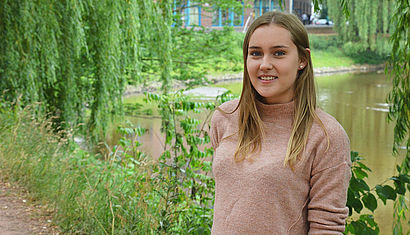 Die 18-jährige Ilka Eßing steht auf einem Weg unter grünen Bäumen, im Hintergrund ist die Bocholter Aa zu erkennen.