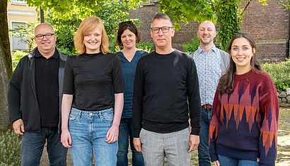 Drei Frauen und drei Männer stehen nebeneinander im Schatten.