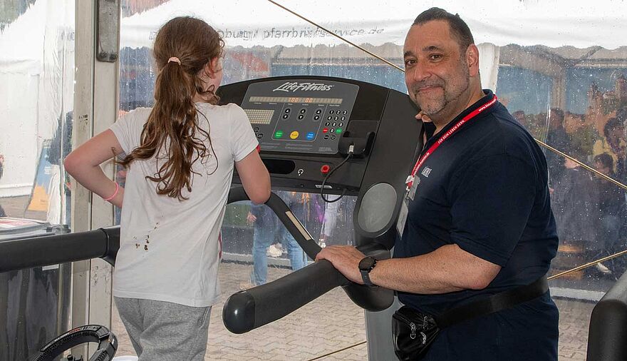 Ein Mann steht an einem Laufband, von hinten ist ein Mädchen zu sehen, das gerade auf dem Laufband ist.