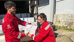 Zwei Helfer in roten Caritas-Jacken übergeben vor einem Transporter Hilfsgüterkisten an Bedürftige.