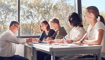 Vier Schüler sitzen an Schreibtischen, davor hockt ein Lehrer.