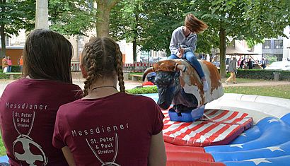 Zwei Mädchen stehen mit dem Rücken zur Kamera und sehen einem dritten Mädchen zu, das auf einem (künstlichen) Bullen reitet.