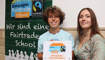 Eine Schülerin und ein Schüler halten eine Fairtrade Plakette in den Händen und stehen vor einem Plakat mit der Aufschrift "Wir sind eine Fairtrade School"