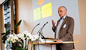 Prof. Dr. Paul Zulehner steht an einem Rednerpult, hinter ihm eine große Leinwand auf der eine Präsentation zu sehen ist.