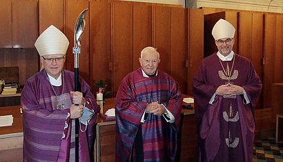 Zwei Bischöfe und ein Priester stehen in Messgewändern in einer Sakristei.