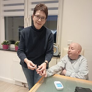 In einem hellen Raum steht eine Frau mit Brille neben einem Tisch und misst mit einer Lanzette den Blutzucker einer sitzenden Person, deren Hand sie hält. Auf dem Tisch liegt ein Blutzuckermessgerät, im Hintergrund sind ein Fenster und kleine Pflanzen zu sehen.