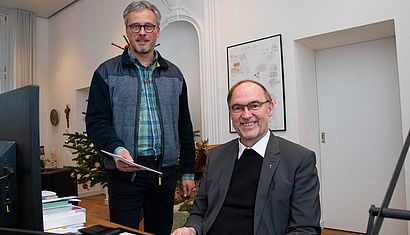 Markus van Berlo steht neben Weihbischof Rolf Lohmann, der am Schreibtisch sitzt.