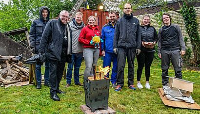 Mehrere Menschen stehen auf einer Wiese, im Vordergrund steht ein Metallkorb mit brennenden Holzscheiten.