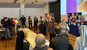 Eine Gruppe von etwa zwanzig Personen steht in einem hellen Seminarraum im Halbkreis. Einige tragen religiöse Ordenstracht. Eine Person präsentiert vor zwei Pinnwänden, die mit vielen farbigen, runden Papierkarten mit einfachen Zeichnungen bedeckt sind. Auf der Leinwand im Hintergrund sind ebenfalls skizzierte Gesichter zu sehen. Tische mit Unterlagen und Getränken stehen im Vordergrund.