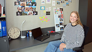 Studentin Lara Sommerfeld sitzt vor dem Schreibtisch in ihrem Zimmer. Im Hintergrund sieht man die Wand, die sie mit zahlreichen Fotos und Stickern dekoriert hat.
