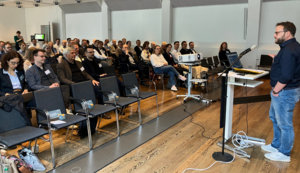 Ein in blaue Jeans und dunkelblauen Hemd gekleideter Mann steht an einem Rednerpult und spricht zu dem vor ihm sitzenden Publikum.