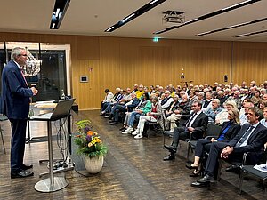 Ein Mann im Anzug steht in einem modernen Saal an einem Pult mit Laptop und Mikrofon und hält einen Vortrag. Vor ihm sitzt ein großes Publikum, überwiegend ältere Erwachsene, die aufmerksam zuhören. Der Raum hat eine Holzvertäfelung, helle Beleuchtung und einen dunklen Holzboden. Neben dem Pult steht ein Blumengesteck. 