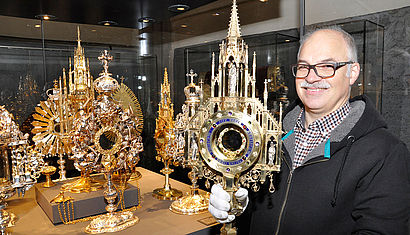 Der Küster Roland van Wegen steht rechts neben einer geöffneten Glasvitrine. Er hält in der rechten Hand eine Monstranz, die gold und silbern glänzt, links stehen weitere Monstranzen.