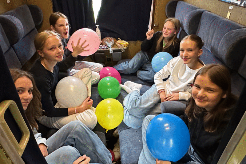 Eine Gruppe von Schülerinnen sitzt mit Luftballons in den Händen in einem Zugabteil.
