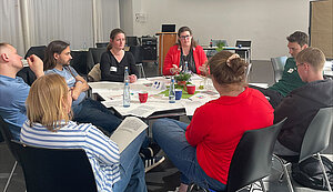 Eine Gruppe von Erwachsenen sitzt an einem runden Tisch in einem Seminarraum und diskutiert gemeinsam. Auf dem Tisch liegen Unterlagen, Notizen und Getränke, während eine Person in roter Jacke spricht und die anderen aufmerksam zuhören.