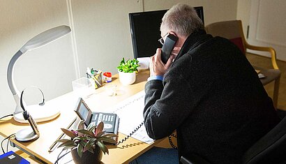 Ein Mann sitzt mit dem Rücken zum Betrachter an einem Schreibtisch und hält einen Telefonhörer ans Ohr.
