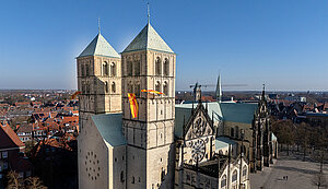 Der St.-Paulus-Dom in Münster an einem sonnigen Tag. Die beiden Türme sind mit gelb-roten Fahnen geschmückt, während sich die Architektur des Doms vor der Kulisse der Stadt und einem klaren blauen Himmel abhebt. Der St.-Paulus-Dom in Münster an einem sonnigen Tag. Die beiden Türme sind mit gelb-roten Fahnen geschmückt, während sich die Architektur des Doms vor der Kulisse der Stadt und einem klaren blauen Himmel abhebt.