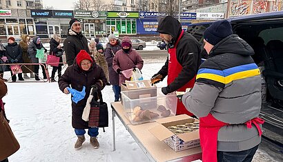 Auf dem Foto wird im Freien auf einer verschneiten Fläche eine Essensausgabe gezeigt. Mehrere Personen stehen in einer Schlange, während an einem Tisch Lebensmittel wie Brot und Konservendosen verteilt werden.