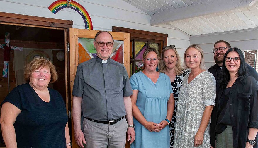 Mehrere Frauen und Männer stehen vor einem Holzgebäude, über dessen Eingang ein kleiner Regenbogen angebracht ist. Mehrere Frauen und Männer stehen vor einem Holzgebäude, über dessen Eingang ein kleiner Regenbogen angebracht ist.