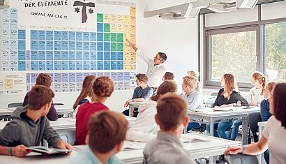 Mehrere Schüler sitzen in einer Klasse und schauen aufmerksam auf den Lehrer, der auf eine bunte Tafel mit dem Periodensystem der Elemente weist.