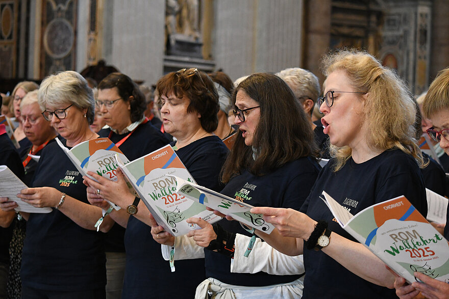 Frauen und Männer singen im Chor mit einheitlichen dunkelblauen Shirts bekleidet und Chorbüchern in der Hand.