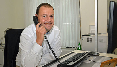 Christian Walbröhl sitzt in einem Büro, vor ihm ist eine Tastatur zu sehen. Mit der rechten Hand hält er sich einen Telefonhörer ans Ohr.