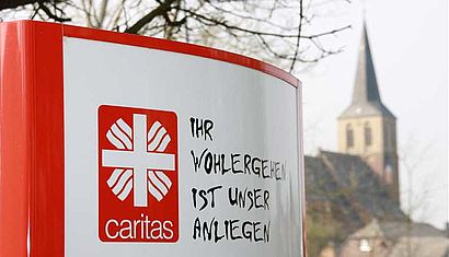 Im Vordergrund ist ein Schild der Caritas zu sehen, im Hintergrund ein Kirchturm.