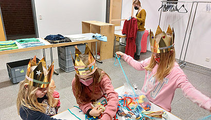 Drei Mädchen mit Maske und Krone suchen sich Gürtel aus. Im Hintergrund hat eine Frau ein Sternsingergewand in der Hand. 