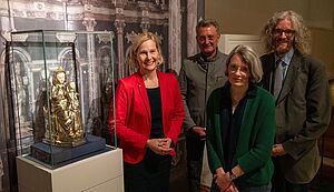 Holger Kempkens, Kuratorin Karin Wermert, Kuratorin Elisabeth Maas und Dompropst Monsignore Joachim Göbel stehen neben einem Glaskasten mit der Reliquienstatue der Thronenden Muttergottes. Im Hintergrund befindet sich die Abbildung des Hochaltarretabels des Domes zu Münster.