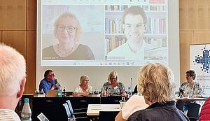 Auf einem Wandmonitor sind Maria Bubenitschek, Moderatorin der Vollversammlung und Vorstandsmitglied, und Referent Dr. Simon Linder von der Universität Tübingen zu sehen. Auf dem Podium davor sitzen der Vorsitzende des Diözesankomitees Ulrich Vollmer, die Vorsitzende Brigitte Lehmann, Moderatorin und Vorstandsmitglied Reinhilde Riesenbeck und Geschäftsführerin Lisa Rotert. Auf einem Wandmonitor sind Maria Bubenitschek, Moderatorin der Vollversammlung und Vorstandsmitglied, und Referent Dr. Simon Linder von der Universität Tübingen zu sehen. Auf dem Podium davor sitzen der Vorsitzende des Diözesankomitees Ulrich Vollmer, die Vorsitzende Brigitte Lehmann, Moderatorin und Vorstandsmitglied Reinhilde Riesenbeck und Geschäftsführerin Lisa Rotert.
